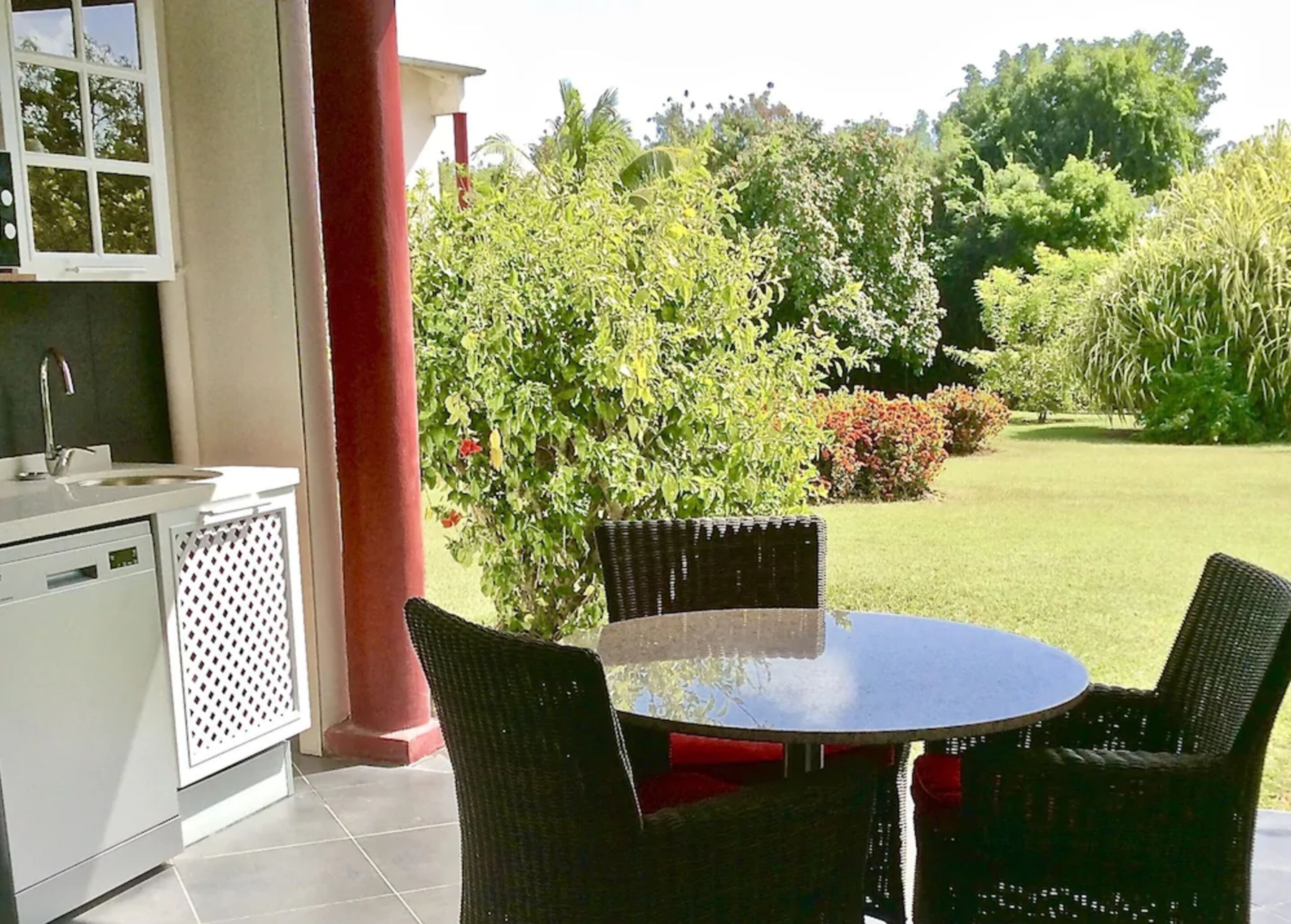 A patio with a round table and wicker chairs, a sink, and a window overlooking a garden with various plants and trees.