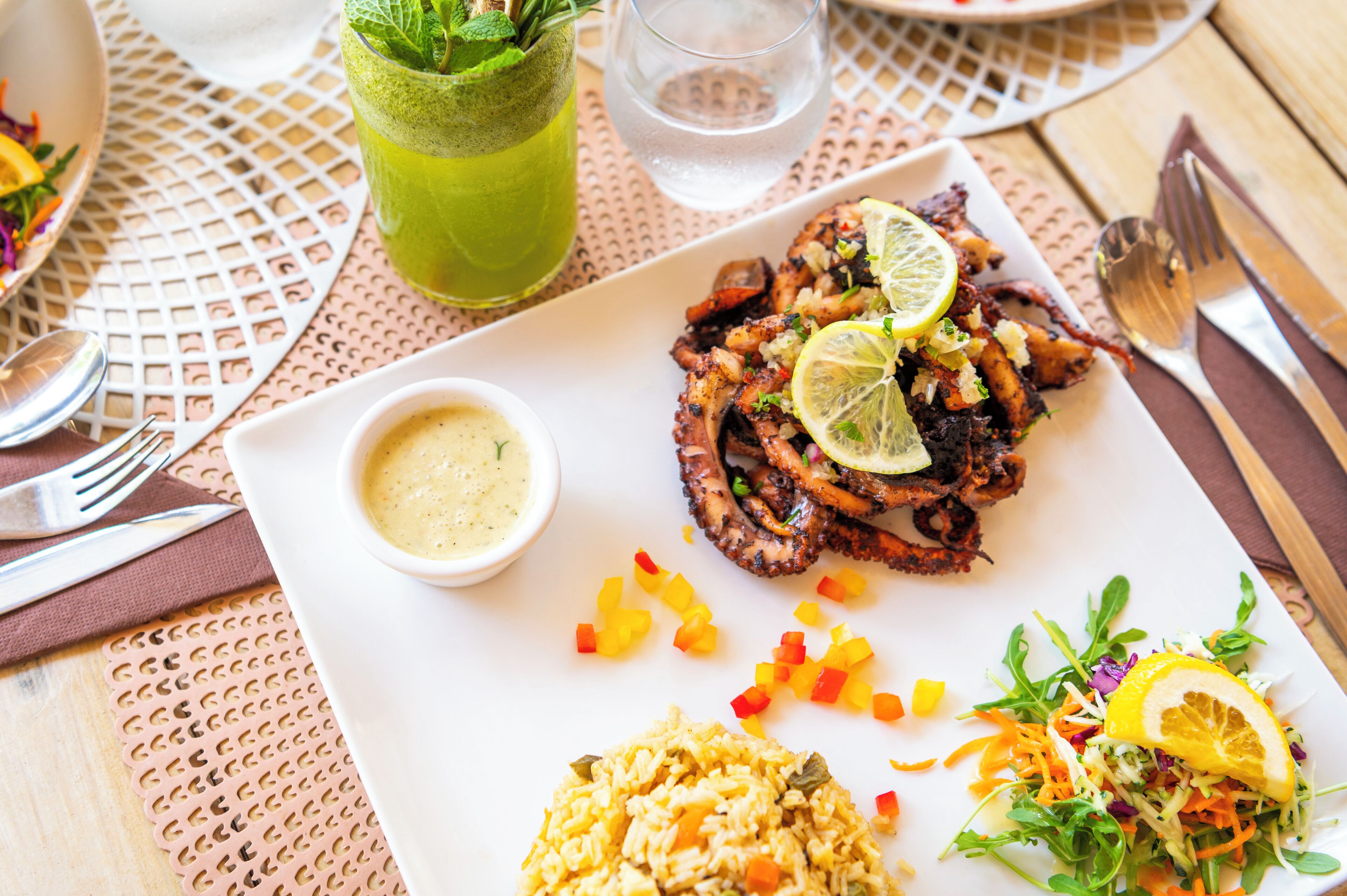 Een eettafel met een bord gegrilde octopus, een schaaltje saus, een glas groene drank en een salade.