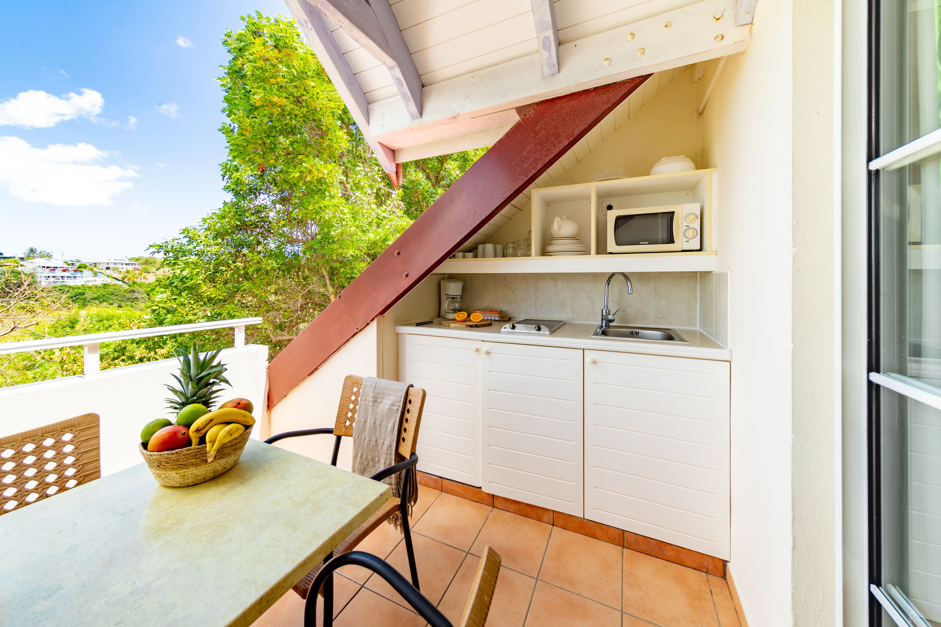 A small kitchen with white cabinets, a microwave, and a sink. There is a table with a fruit basket and a chair. A wooden staircase leads to an outdoor area with a view of greenery.