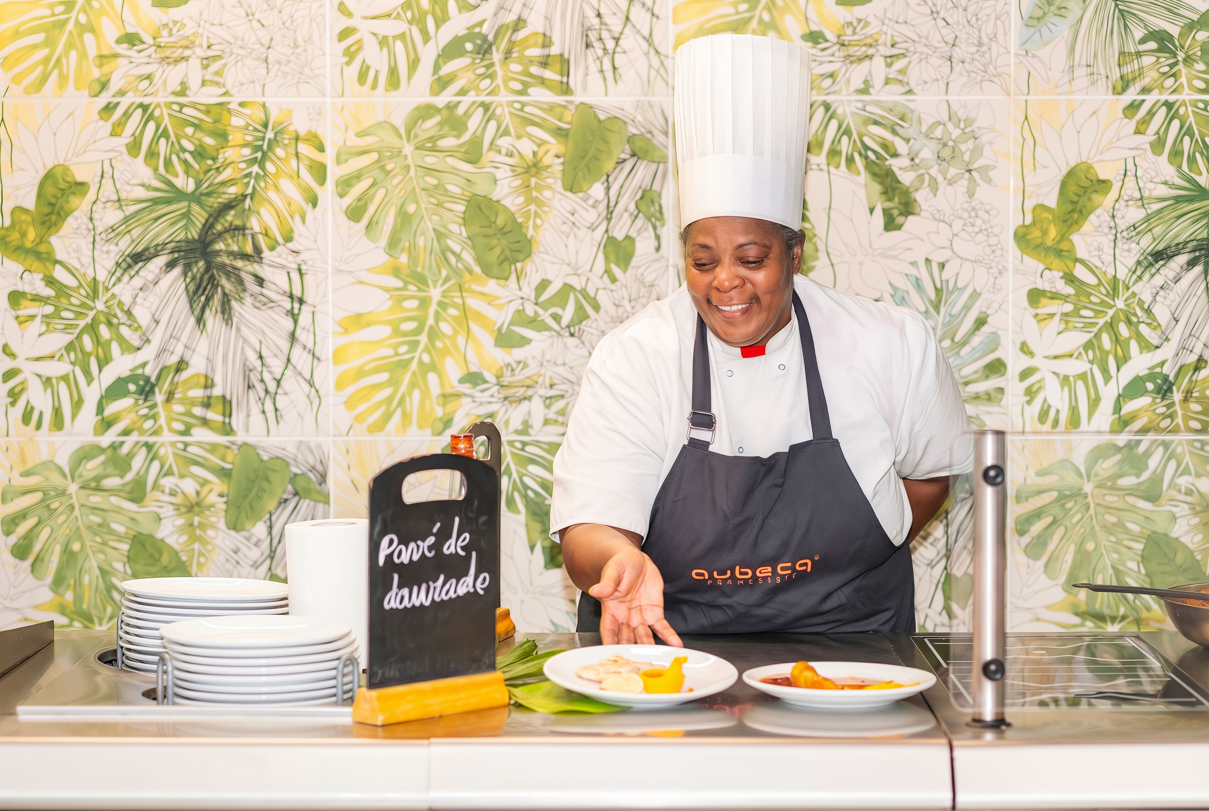 A chef in a white chef's hat and black apron, preparing food at a counter with a tropical leaf-patterned backsplash.