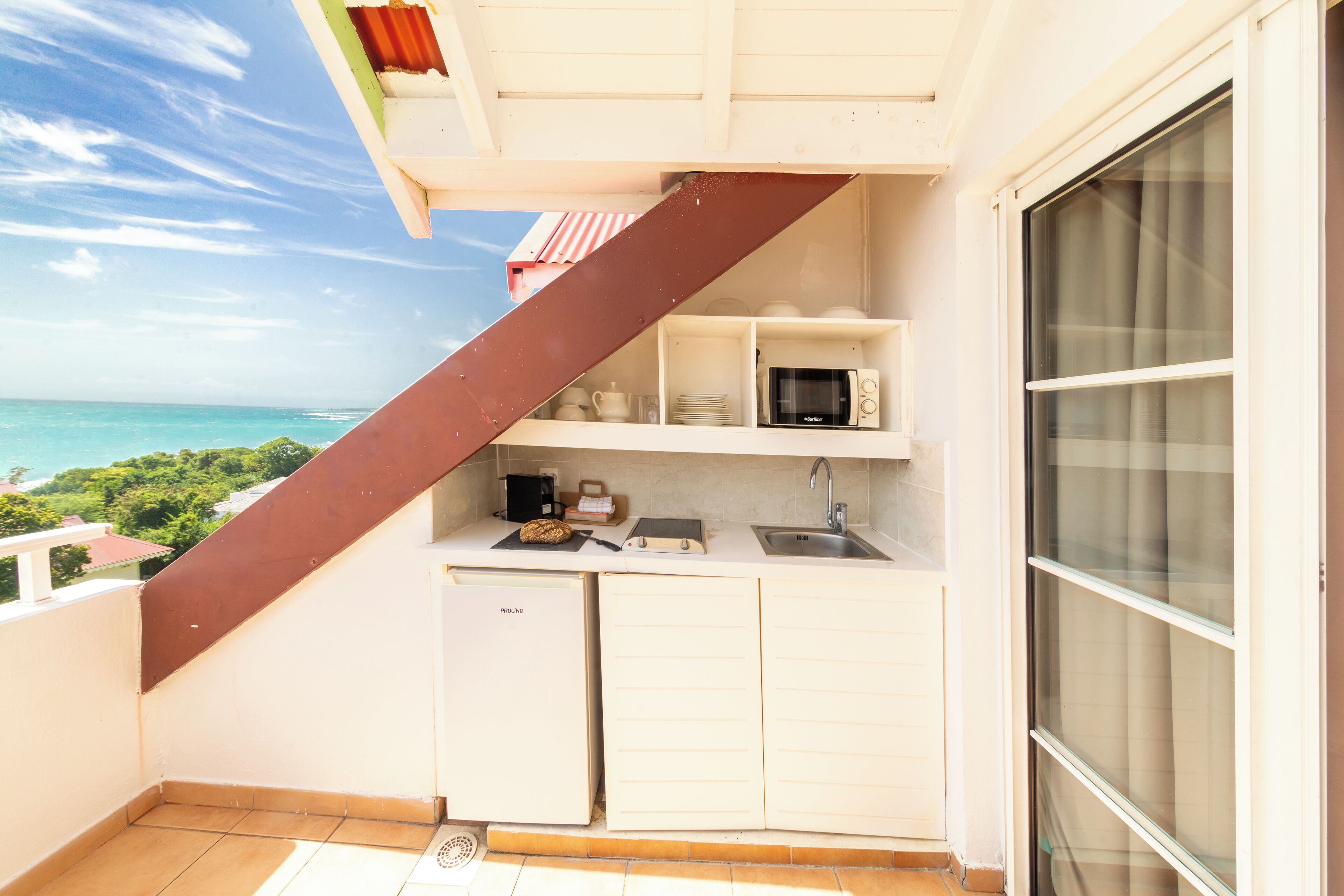 A compact kitchen with a microwave, sink, and a small refrigerator. It features a balcony with a view of the ocean.