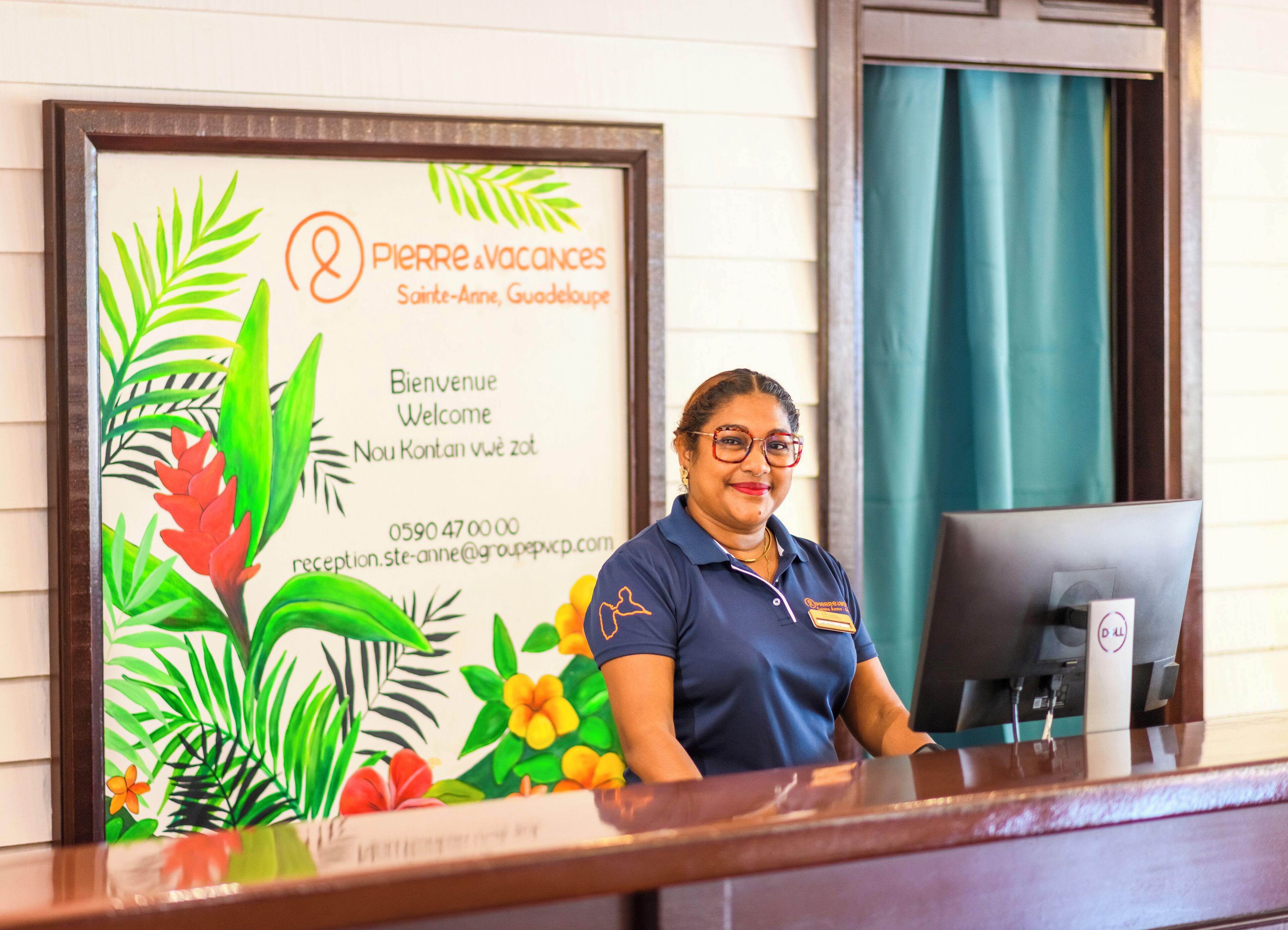A reception desk with a framed welcome sign featuring tropical plants and contact information for a hotel in Sainte-Anne, Guadeloupe.