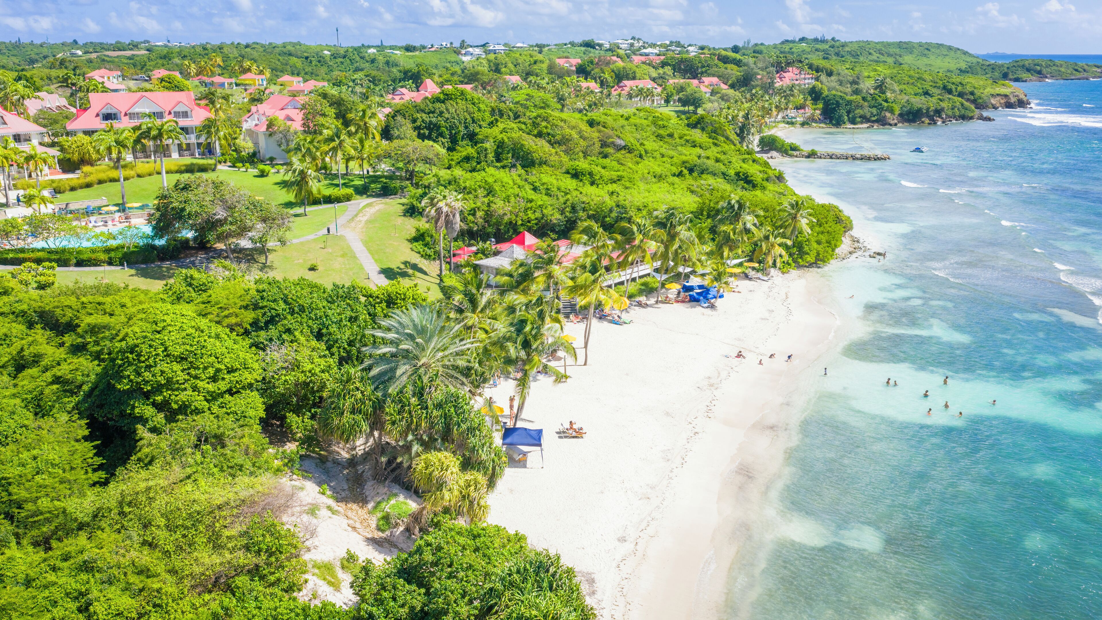 Un complejo turístico frente al mar, con una playa, abundante vegetación y edificios con techos rojos.