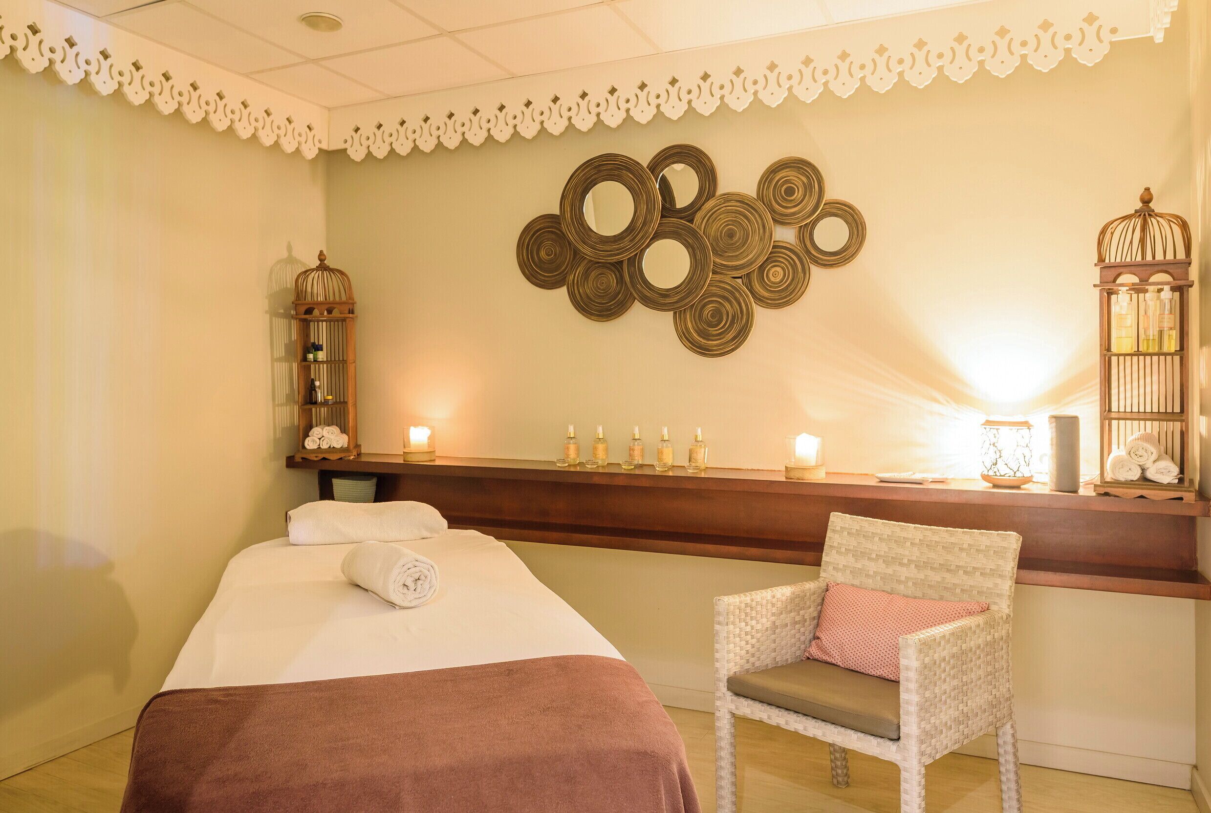 A room with a bed, a chair, a wall with circular mirrors, and a shelf with bottles and a lamp.
