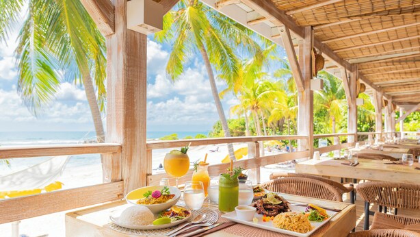 A beachside restaurant with outdoor seating, a view of the ocean, and a variety of dishes on the table. - Village St Anne Pierre & Vacances (Sainte-Anne)