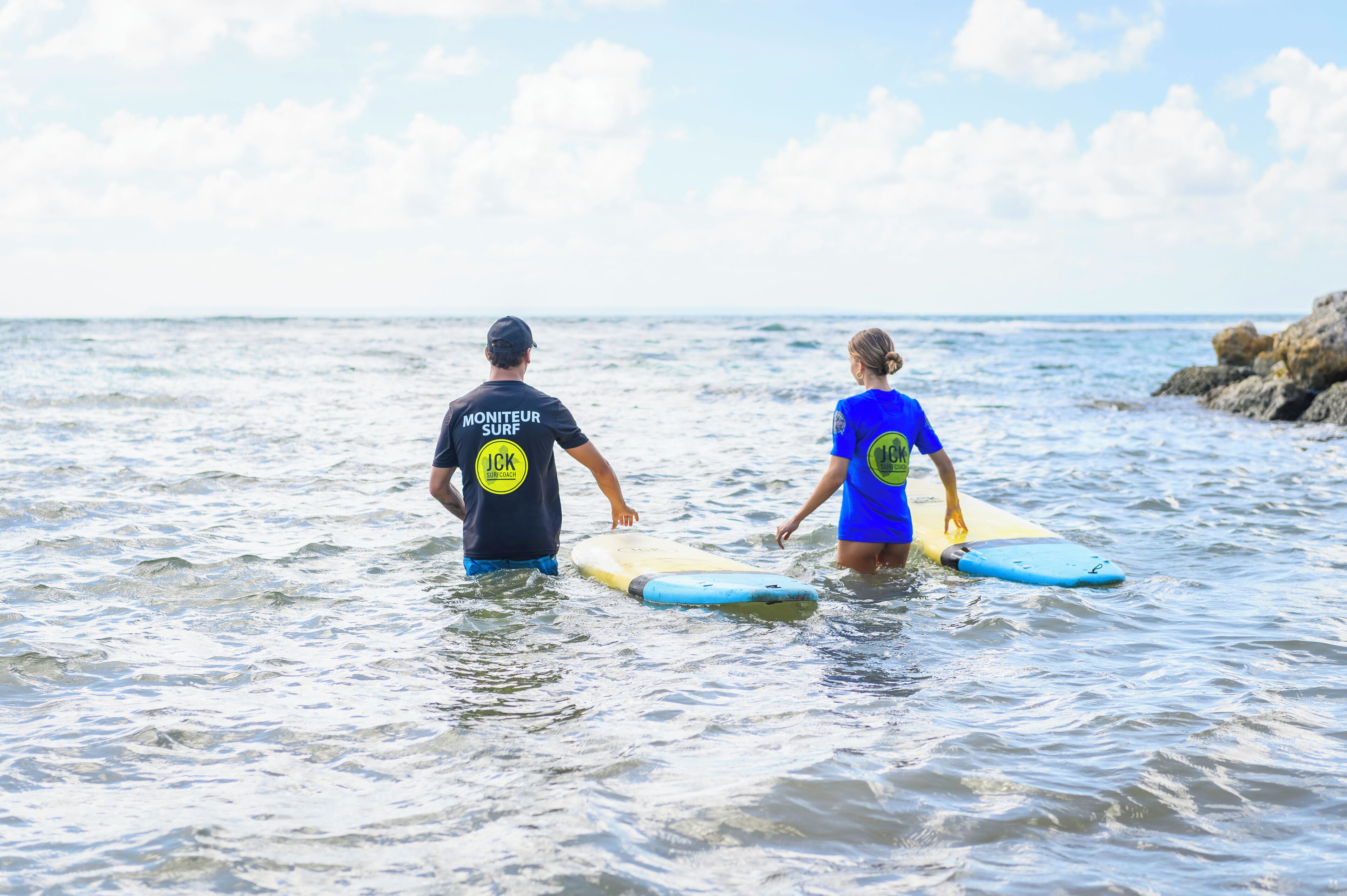 Two people in the water with surfboards, wearing t-shirts with logos.