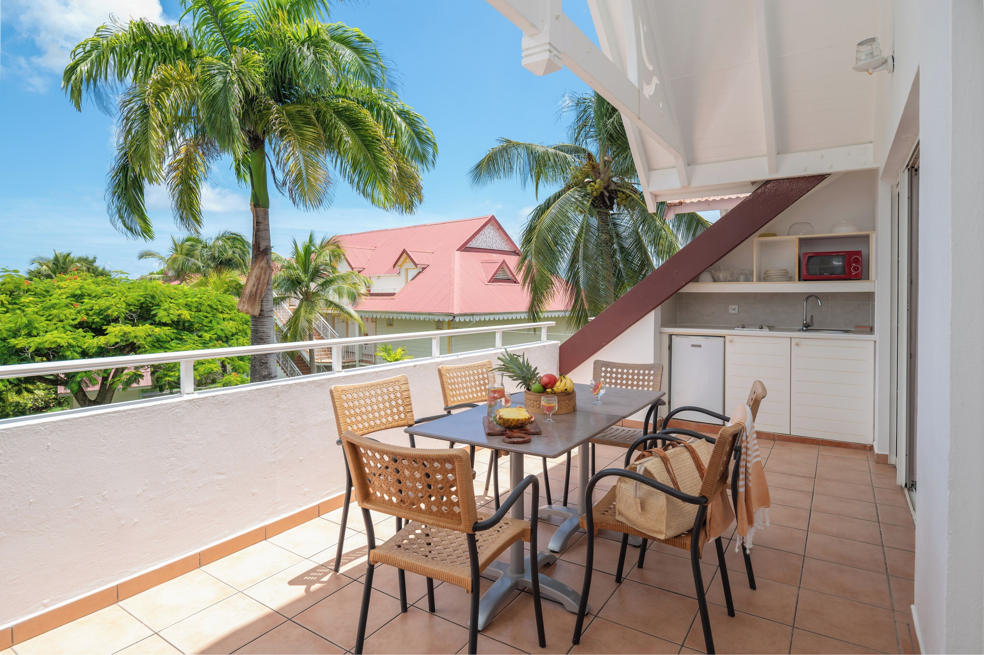 A balcony with a table set for four, surrounded by wicker chairs, with a view of palm trees and a building in the background.
