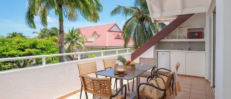 A balcony with a table set for four, surrounded by wicker chairs, with a view of palm trees and a building in the background.