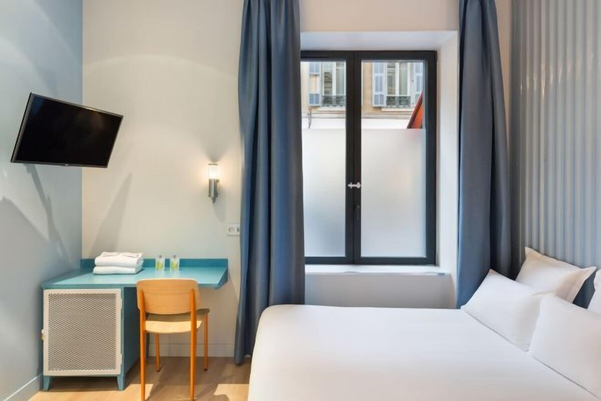 Habitación de hotel con cama, escritorio y silla, televisor montado en la pared y ventana con cortinas.