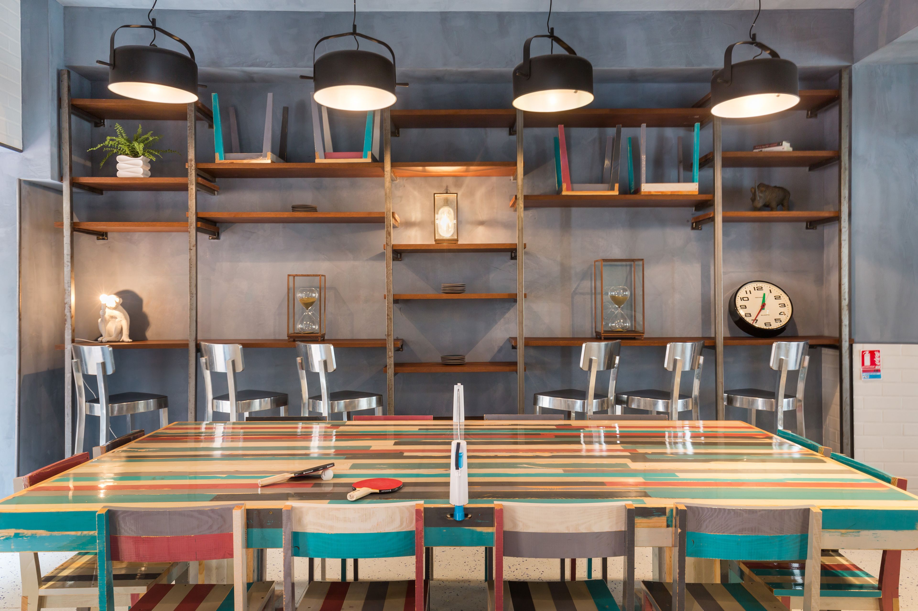 A dining area with a long table, chairs, and overhead lighting. Shelves with books and decorative items are visible.