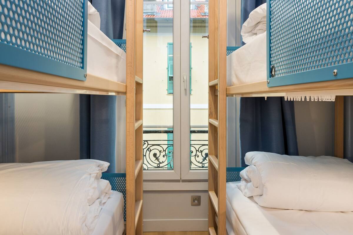 Une petite pièce avec deux lits superposés, une fenêtre avec des rideaux et un balcon doté d’une rambarde en métal.