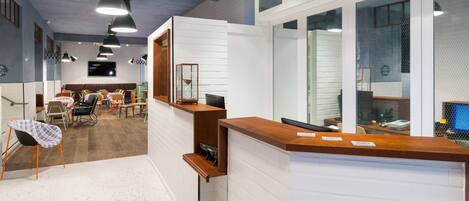 A modern reception area with a wooden reception desk, a seating area with tables and chairs, and a large window with a view of the interior.