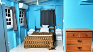 A bedroom with a bed, a wooden dresser, a window with curtains, and a wall-mounted air conditioner. - Chez Ophelia Cottage Apartments (Roseau)