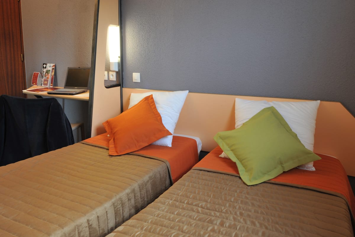 Een hotelkamer met twee bedden, elk met een ander kleurend kussen, een bureau met een laptop en een spiegel aan de muur.