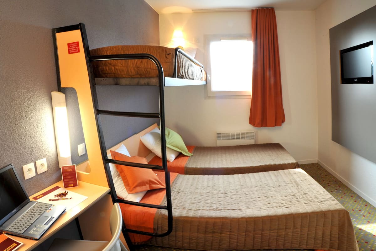 Een kleine hotelkamer met een stapelbed, een bureau met een laptop, een televisie en een raam met gordijnen.