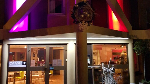 Ein Hotel mit einem gut sichtbaren A&H Schild, einem Weihnachtsgesteck und einem Eingang, an dem ein Schild "CANAL+ LE BOUQUET" angebracht ist.
