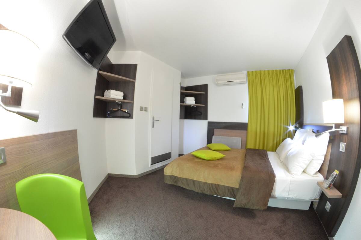 A compact hotel room with a bed, a TV mounted on the wall, a small seating area, and a window with curtains.