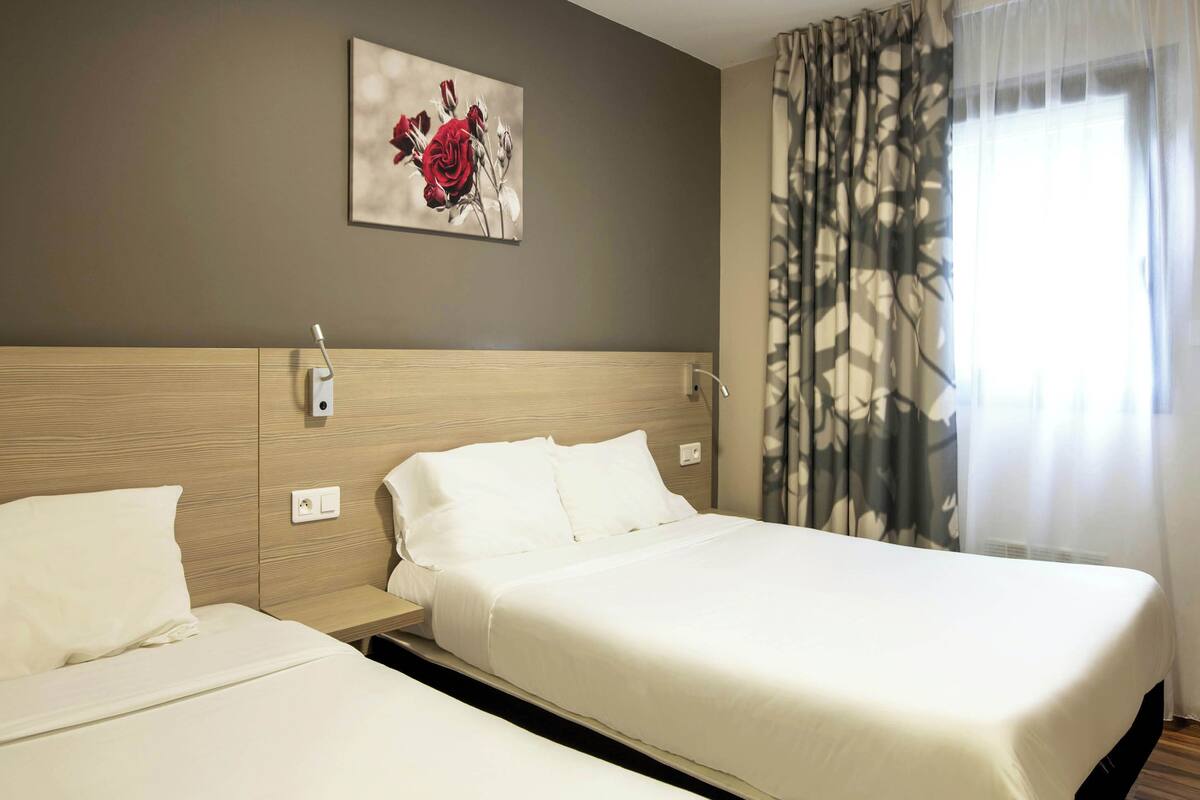 Hotellihuone, jossa on kaksi sänkyä, puinen pääty, seinään kiinnitetty valaisin ja ikkuna, jossa on läpikuultavat verhot.