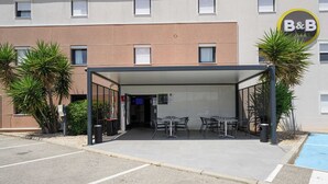 Un área de estar al aire libre techada con mesas y sillas, un edificio con el logo de B&B, y una piscina al fondo.