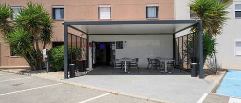 A covered outdoor seating area with tables and chairs, a building with a B&B logo, and a pool in the background.