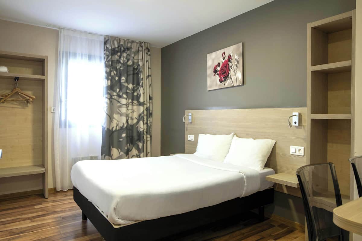 Hotellihuone, jossa on sänky, yöpöytä, tuoli, vaatekaappi ja taulu seinällä.