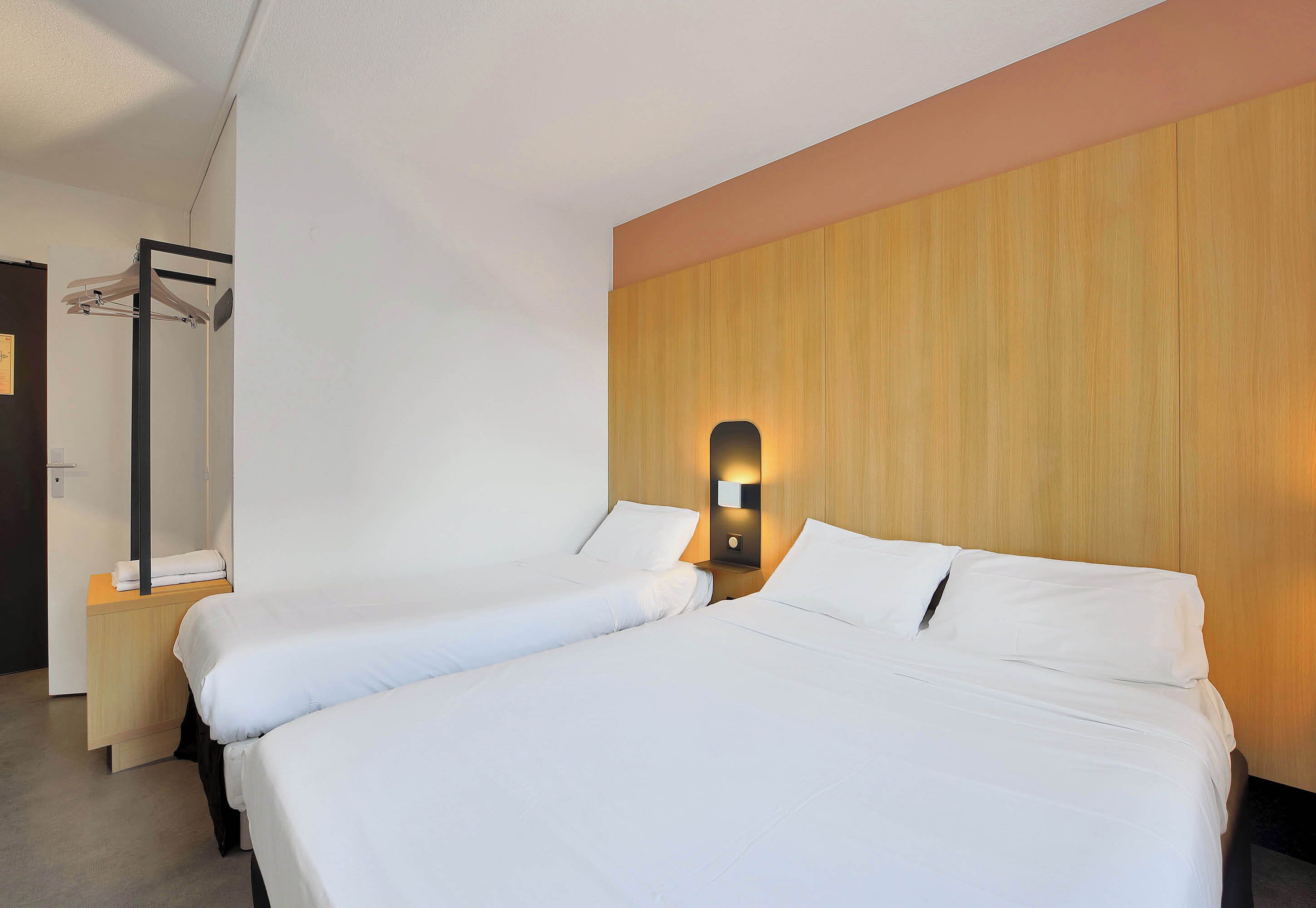 A hotel room with two beds, a wooden headboard, a nightstand, and a wall-mounted light fixture.