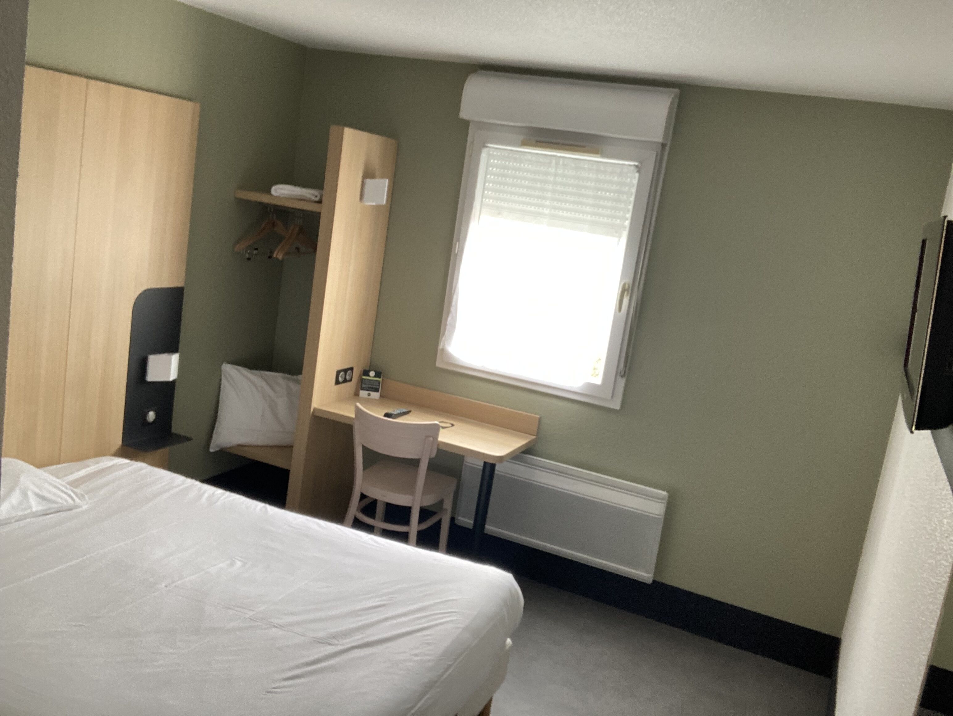 Hotellihuone, jossa on sänky, työpöytä, tuoli, vaatekaappi ja ikkuna, jossa on sälekaihtimet.