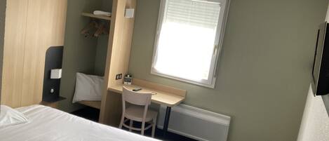 A hotel room with a bed, a desk, a chair, a wardrobe, and a window with blinds.