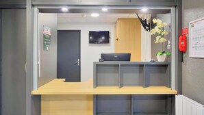 A reception desk with a flat-screen TV above it, a wooden cabinet to the right, and a red fire alarm on the wall.