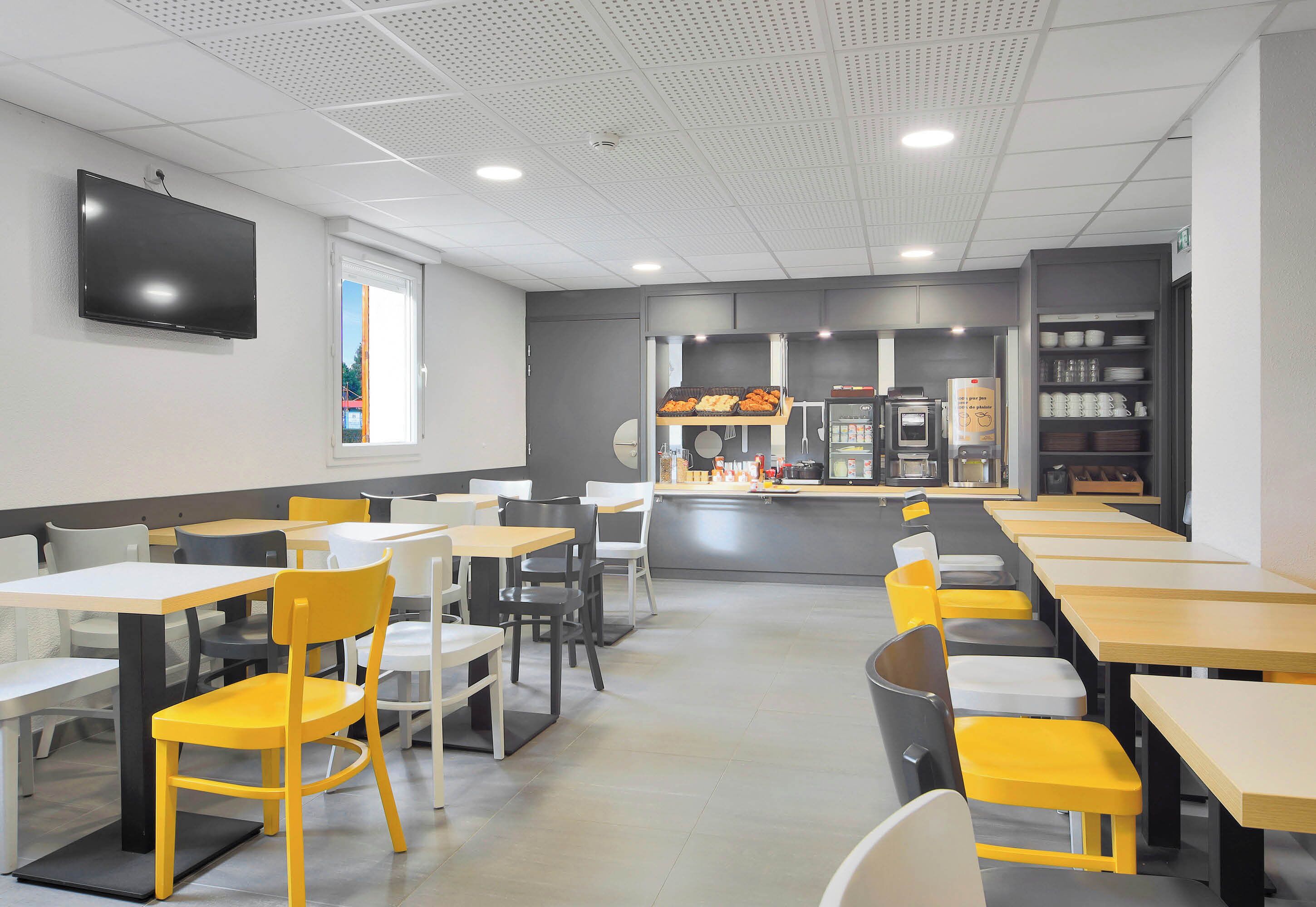 A modern dining area with multiple tables and chairs, a kitchen counter, and a flat-screen TV mounted on the wall.