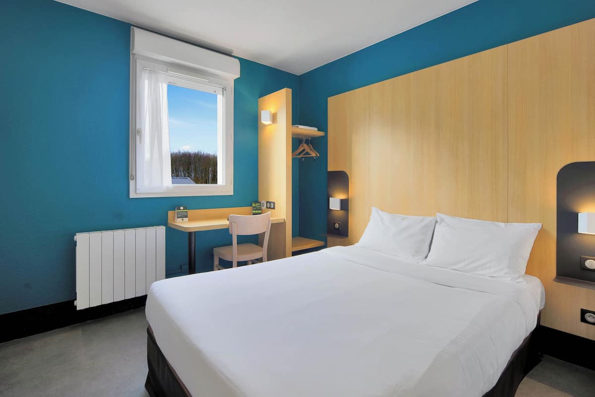 A hotel room with a large bed, a desk with a chair, a window, and a wooden wardrobe.