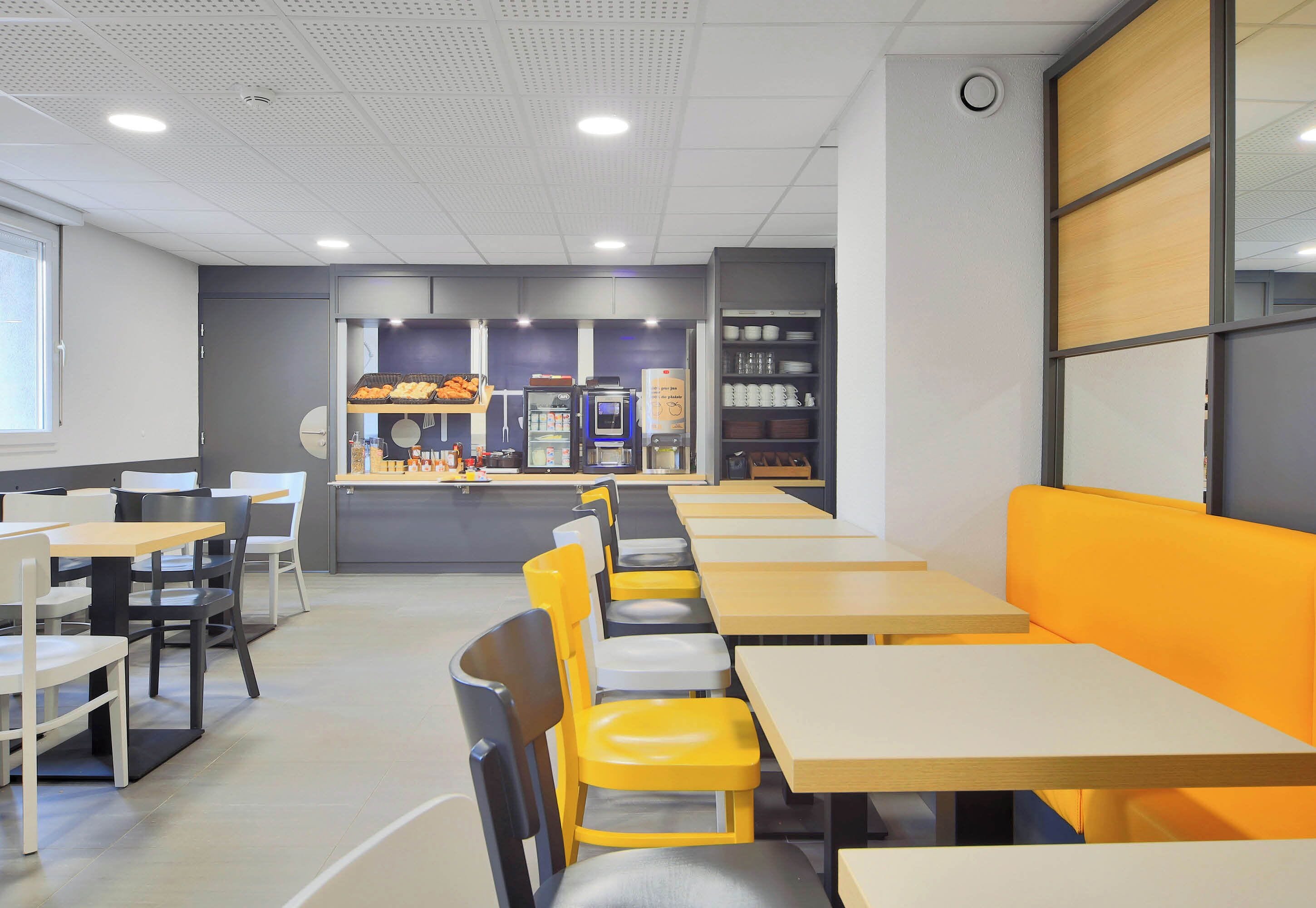 Uma cafeteria moderna com mesas e cadeiras compridas, um balcão com opções de comida e uma área de estar com um sofá amarelo.