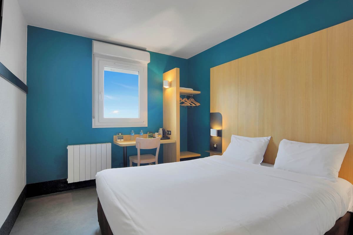 A hotel room with a large bed, a desk with a chair, a window, and a wooden wardrobe.
