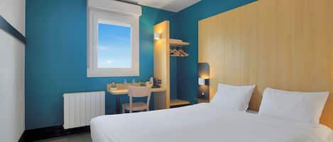 A hotel room with a large bed, a desk with a chair, a window, and a wooden wardrobe.