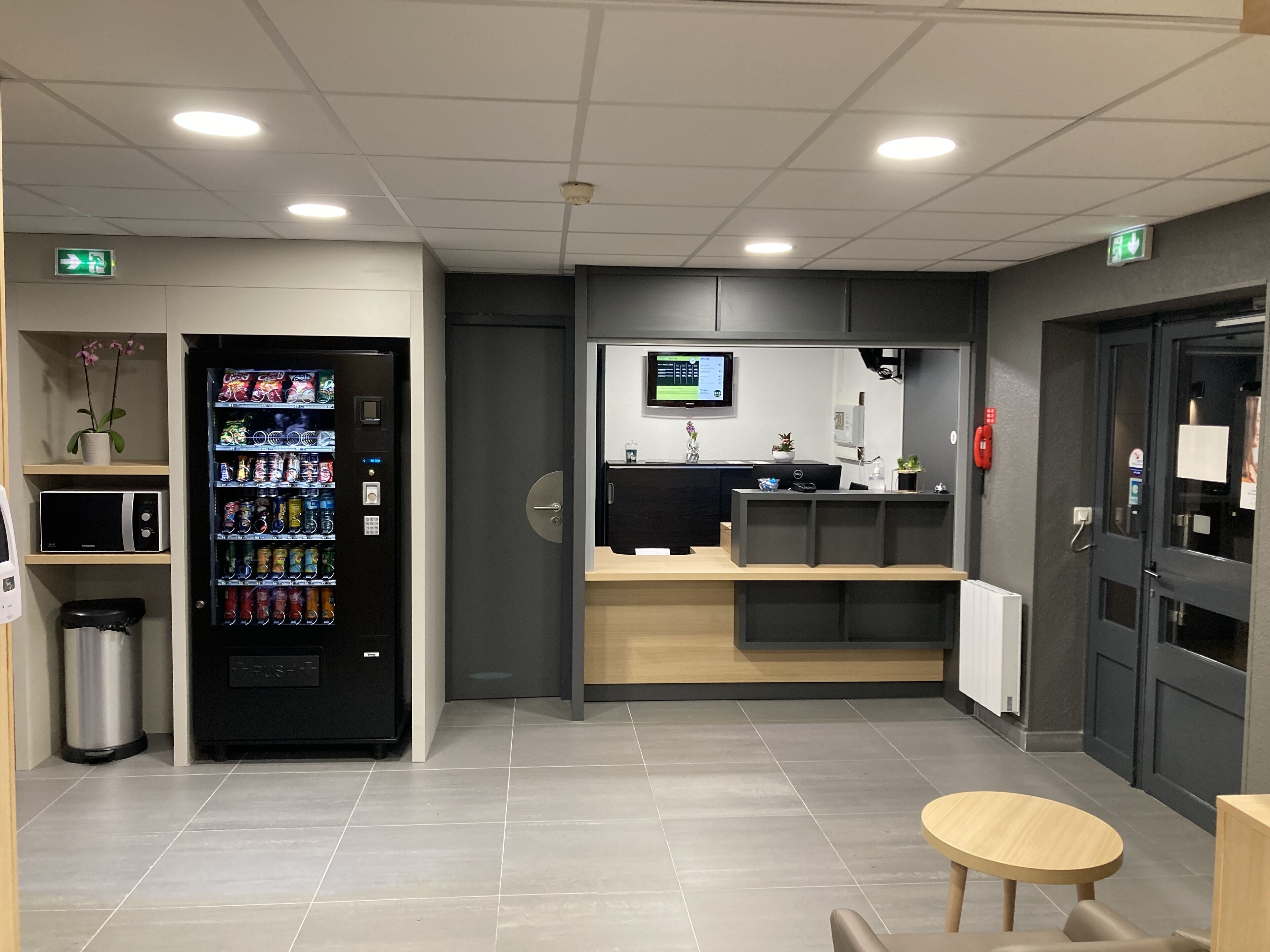 En modern hotellobby med en försäljningsautomat, en receptionsdisk, en digital skärm och en liten sittgrupp.