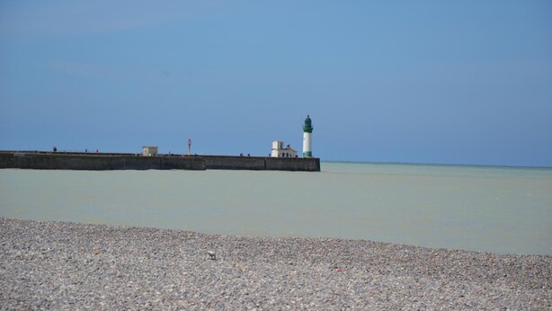 A pebbled beach with a lighthouse and a pier extending into calm waters.