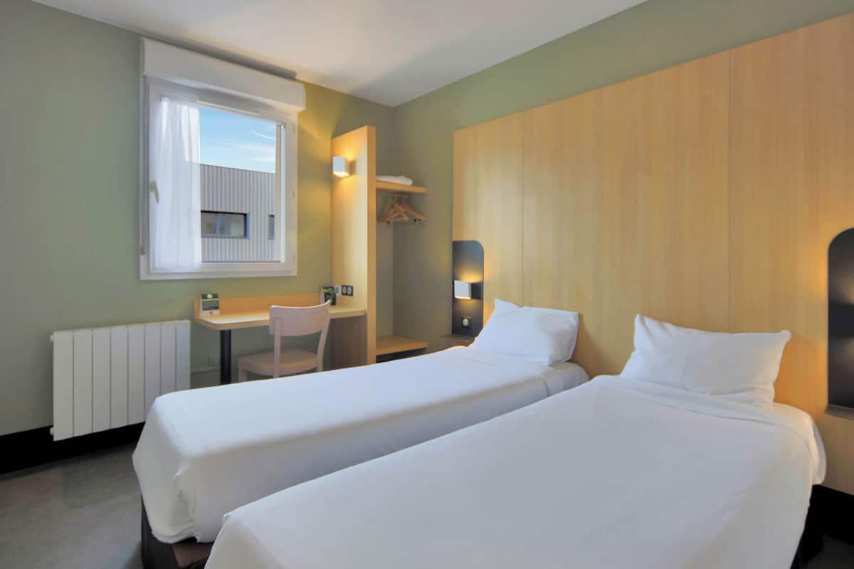 A hotel room with two single beds, a desk with a chair, a small table, a window with curtains, and a wall-mounted shelf with a hanging garment.