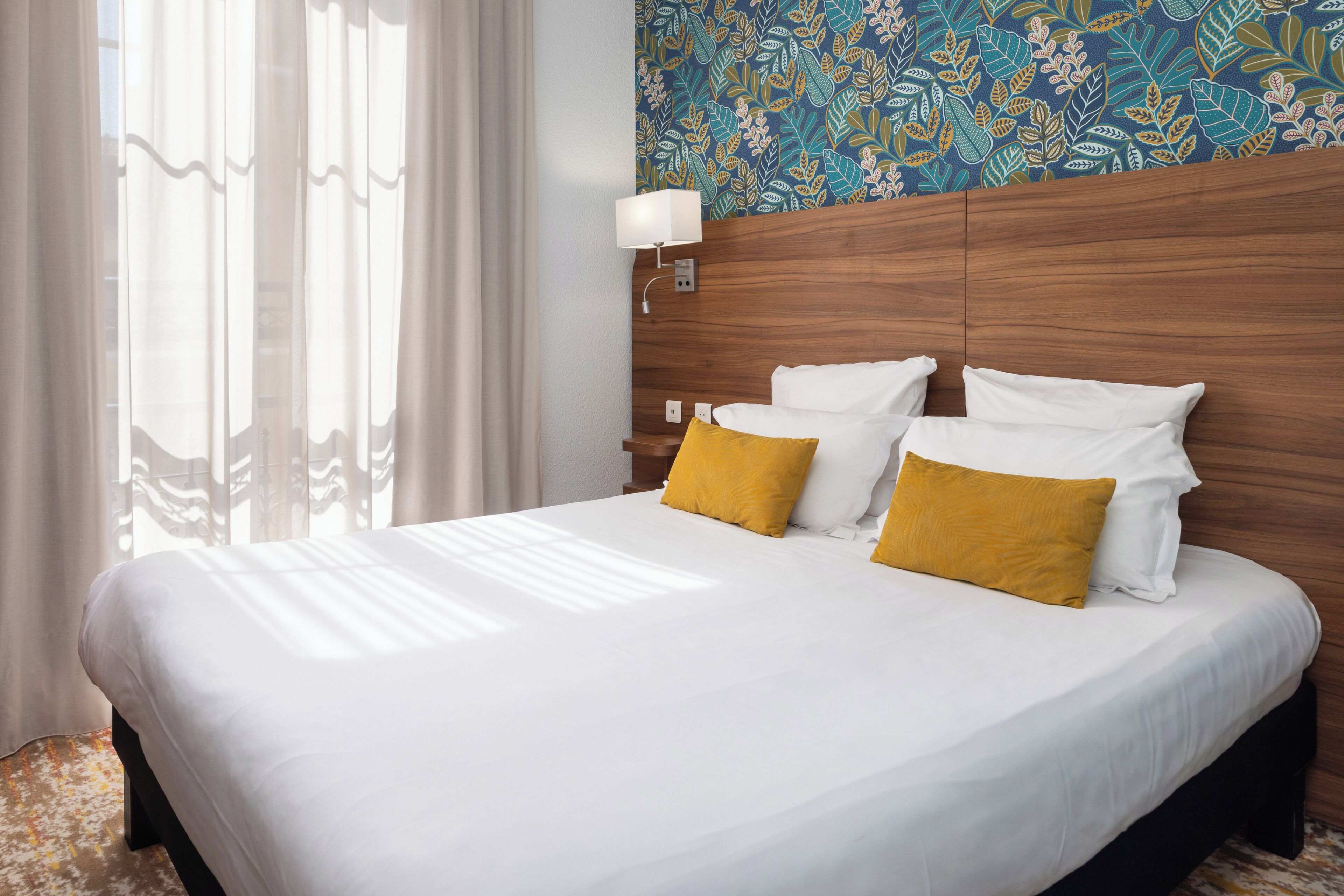 A hotel room with a large bed, white bedding, a wooden headboard, a wall-mounted lamp, and a floral-patterned wallpaper.