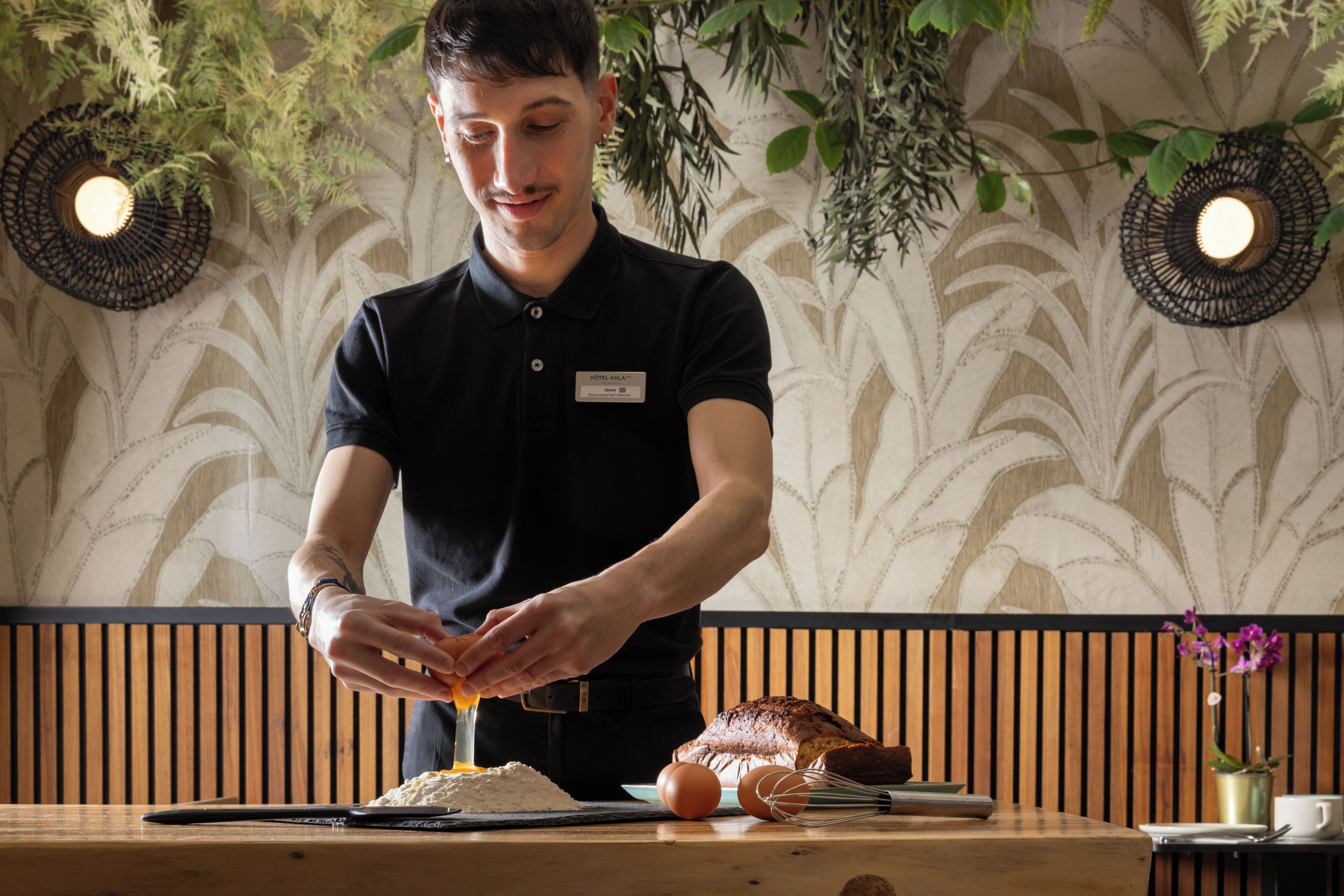 A person preparing food at a wooden table with a patterned wall and decorative lighting in the background.