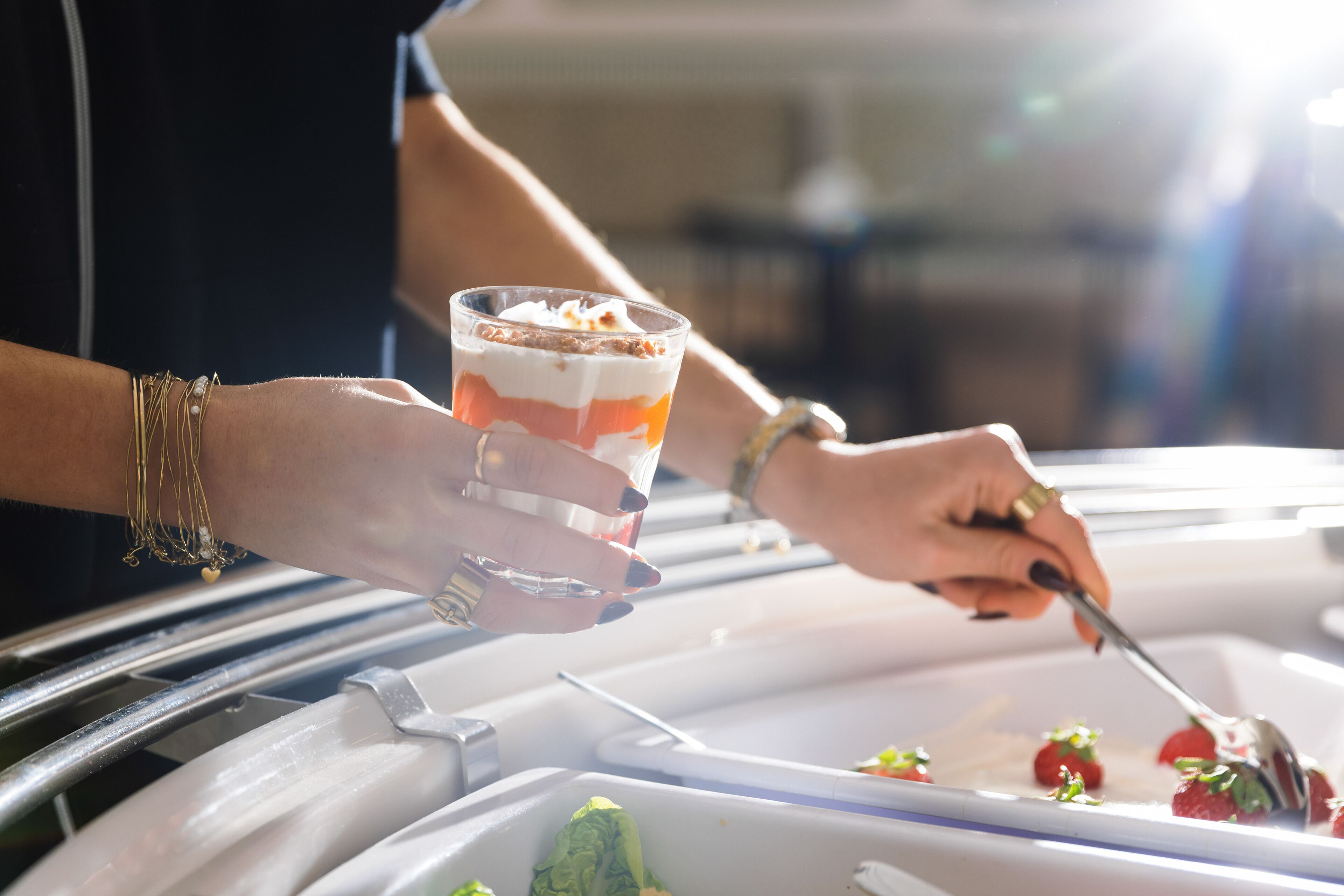 Una persona sosteniendo un postre en capas dentro de un vaso, con una bandeja de fresas y un plato de vegetales verdes al fondo.