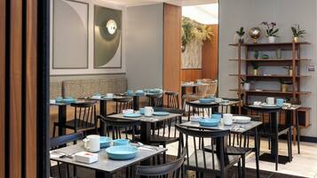 A restaurant with multiple tables and chairs, a wooden shelf with decorative items, and a wall with vertical stripes and a round light fixture.