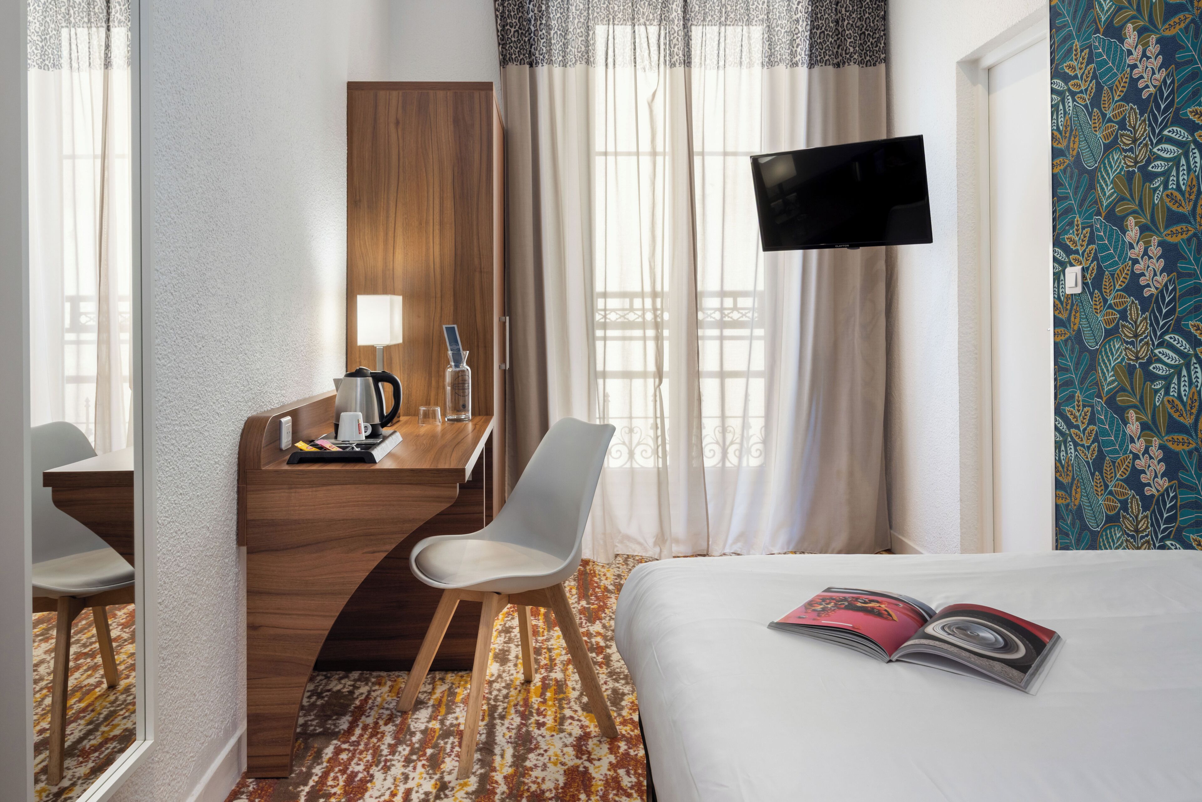 A hotel room with a bed, a desk with a chair, a TV, and a window with curtains.