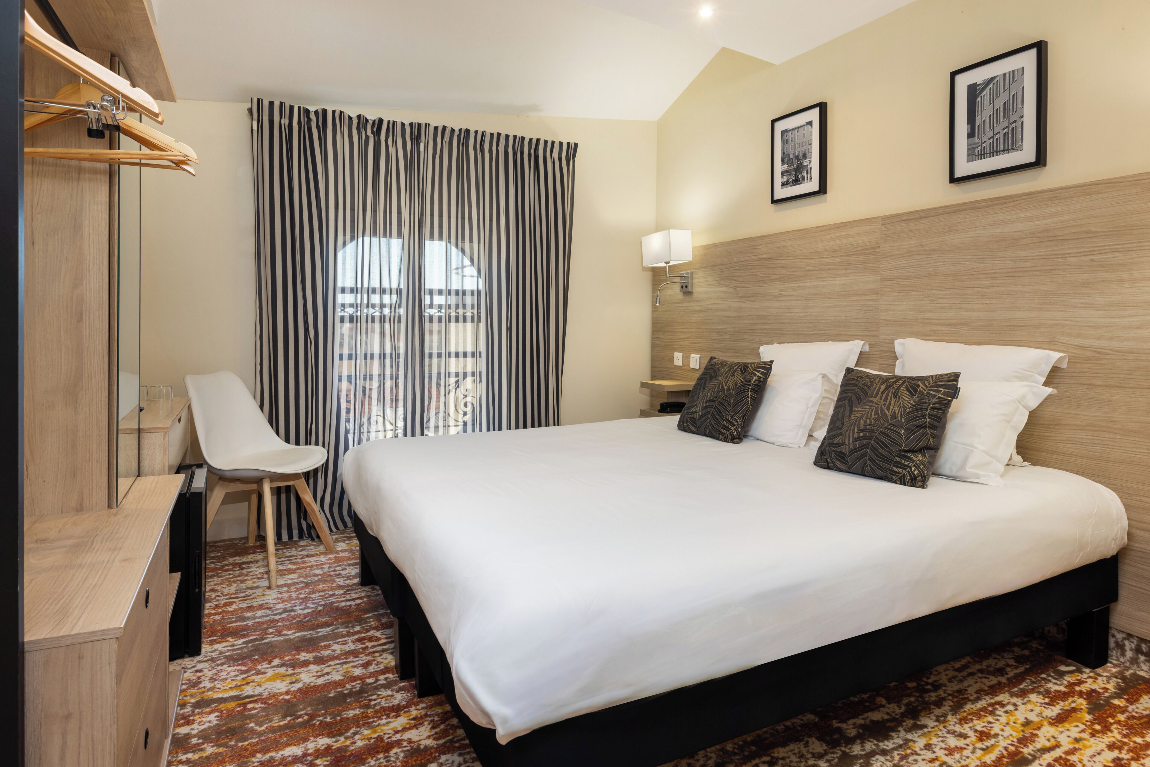 A hotel room with a bed, a desk, a chair, a window with curtains, and a wall-mounted lamp.
