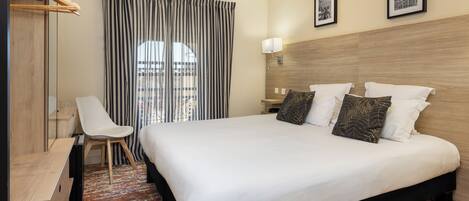 A hotel room with a bed, a desk, a chair, a window with curtains, and a wall-mounted lamp.
