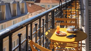 Ein gelber Tisch mit einem Glas Orangensaft, einer Tasse und einem Teller Essen auf einem Balkon mit schwarzem Geländer.