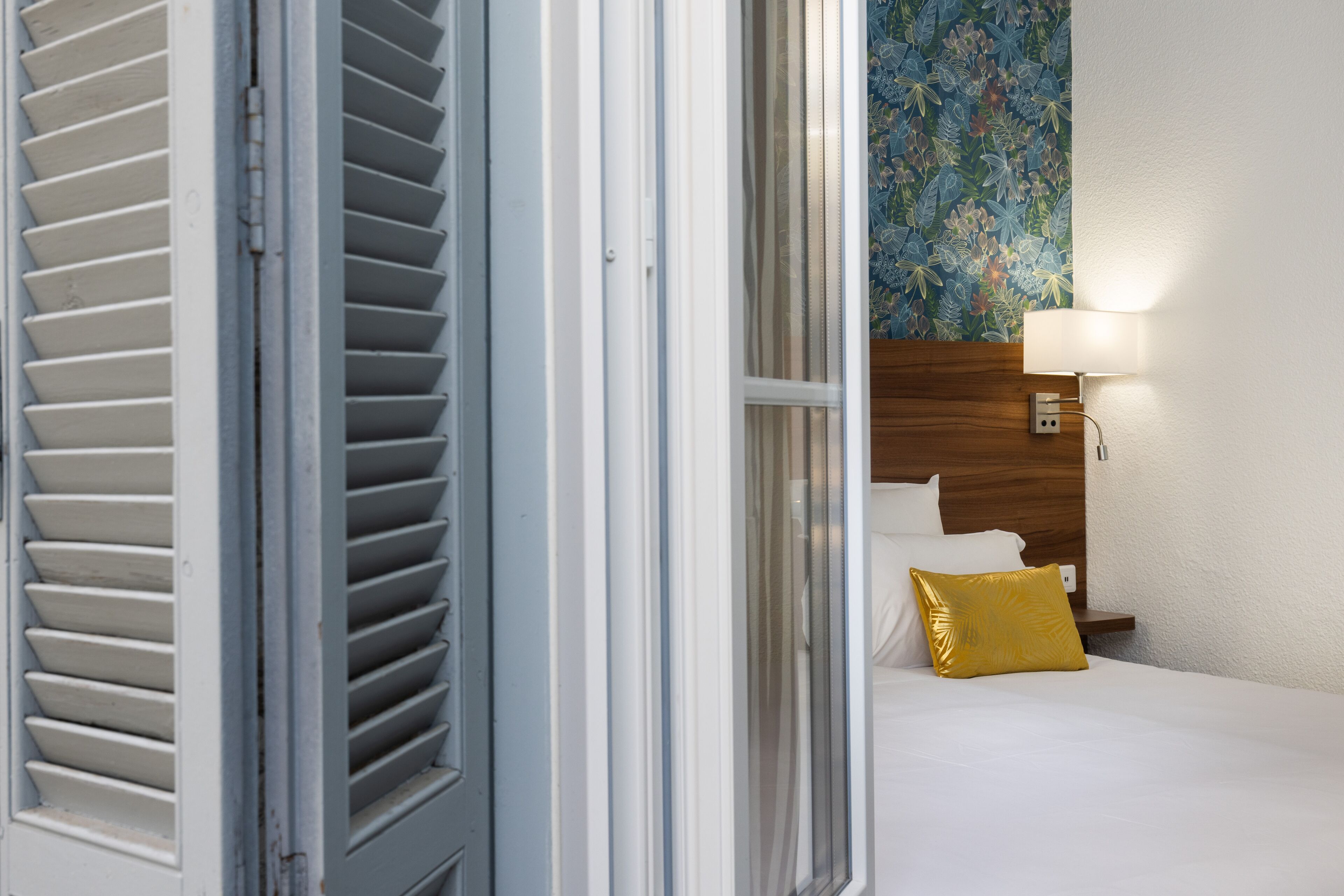 A hotel room with a bed, a nightstand, a lamp, and a window with blinds.