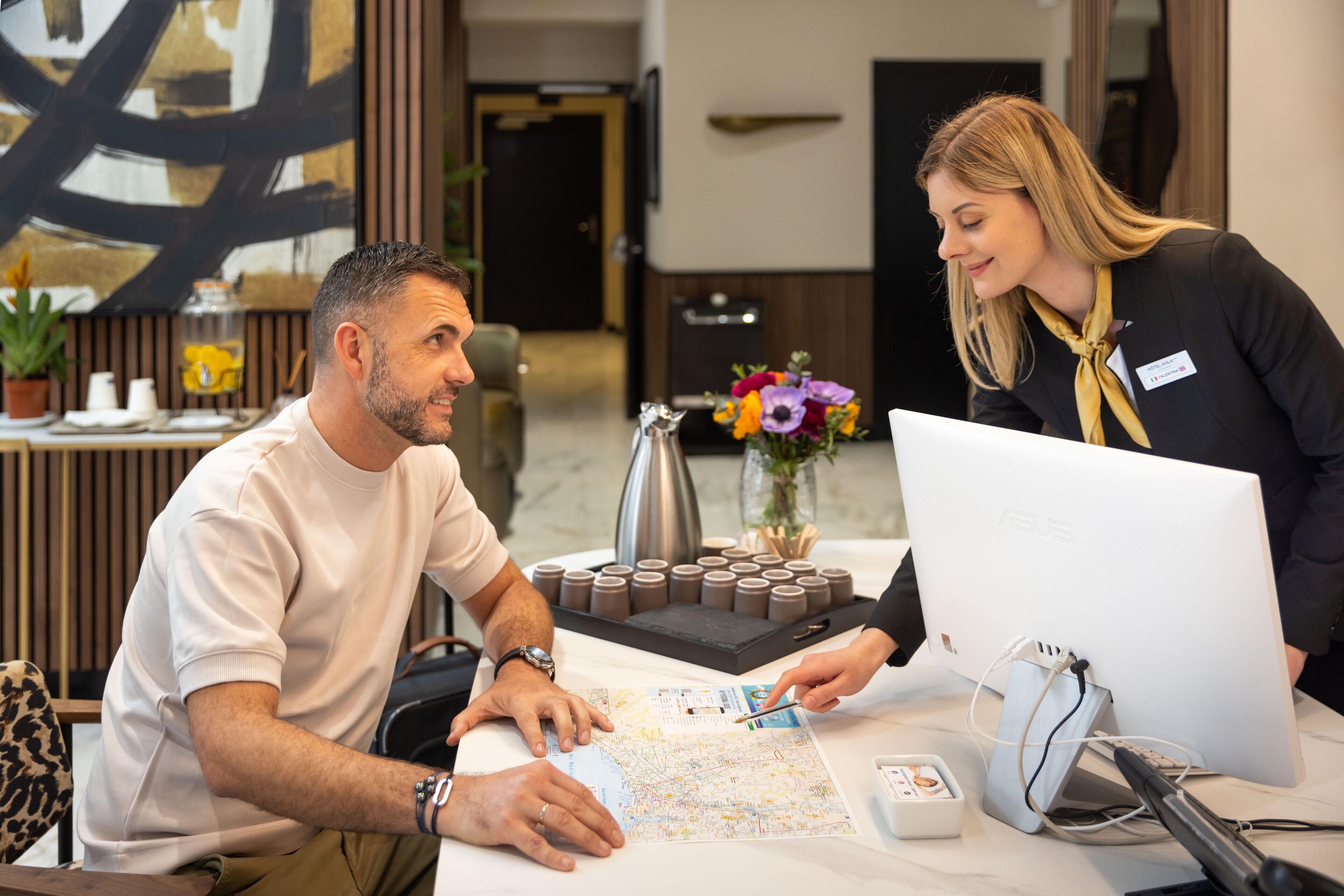 A man and a woman at a reception desk with a map, a computer, and a coffee pot.