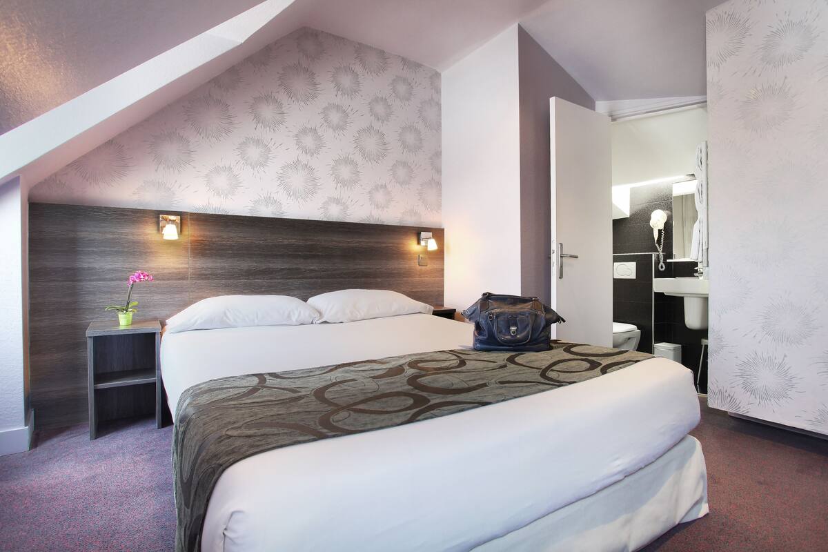 Ein Hotelzimmer mit Bett, Nachttisch, Lampe und einem durchgegangenen Türrahmen einsehbaren Badezimmer.