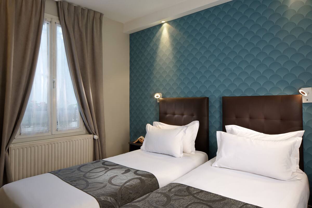 Ein Hotelzimmer mit zwei Betten, einem Fenster mit Vorhängen, einer Wand mit Strukturmuster und einem Nachttisch mit Lampe.