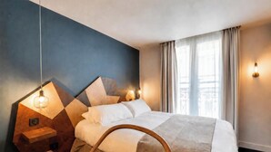 Una habitación de hotel moderna con un cabecero de diseño geométrico, una mesita de noche de madera, una cama con sábanas blancas y vistas a la ciudad a través de amplias ventanas.